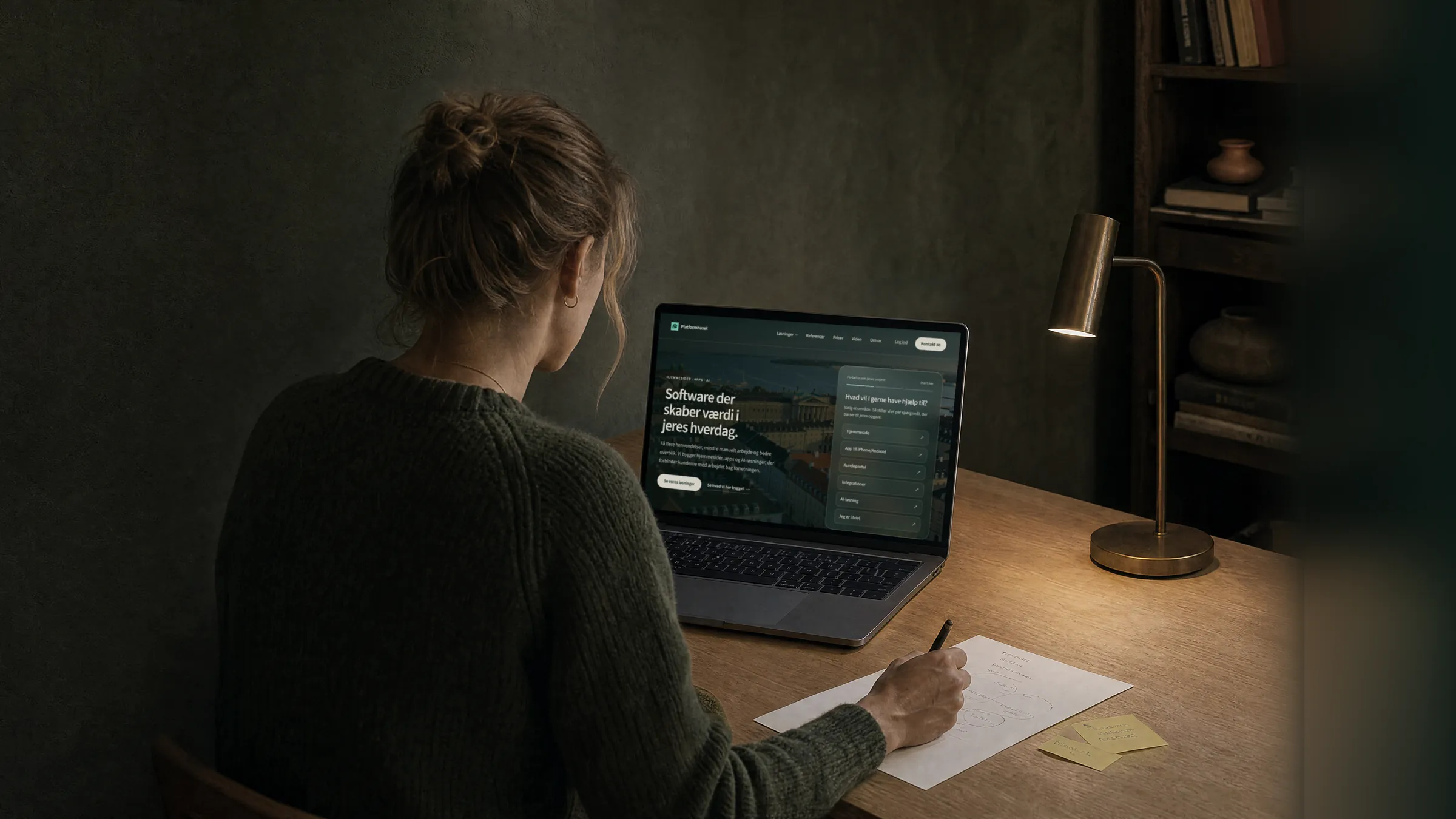 En kvinde i mørkegrøn strik sidder ved et skrivebord og gennemgår en hjemmeside på sin bærbare computer, mens hun noterer med blyant på et ark papir.